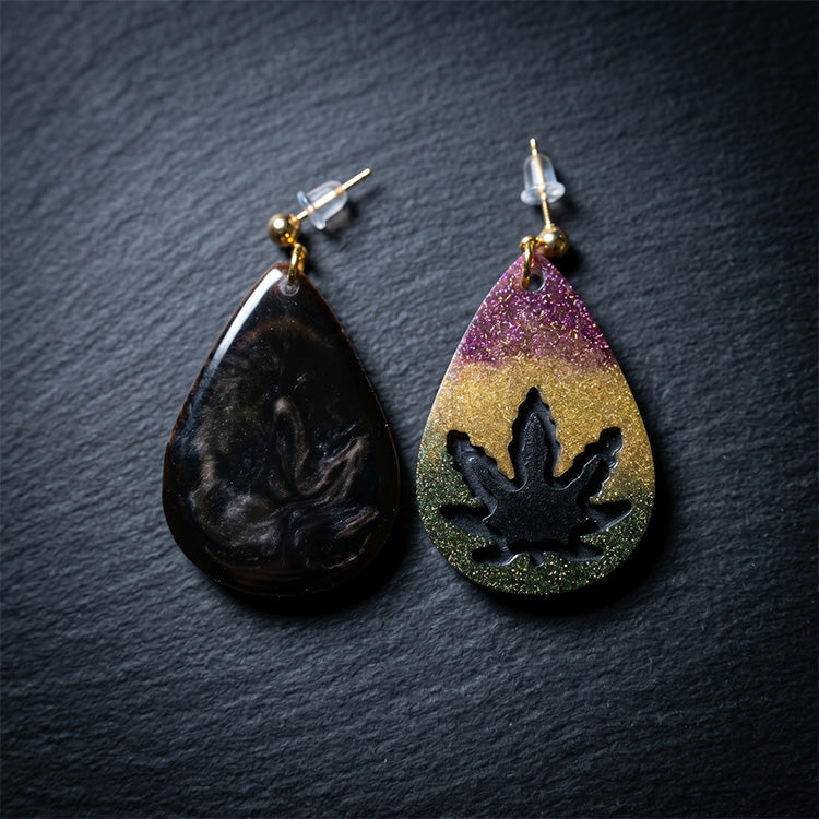 Top-down view of asymmetrical teardrop earrings in gold-purple gradient and dark marbled resin on black slate.