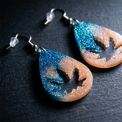 Blue and peach holographic glitter resin earrings with a leaf-shaped cutout design on a dark surface.