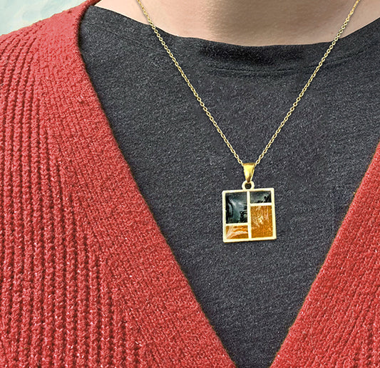 A person wearing a textured red sweater and black shirt displays a gold necklace with a square pendant divided into four sections, each with a different color or design.