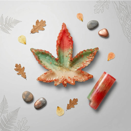 Overhead view of a cannabis leaf resin ashtray and lighter sleeve with a red-to-green gradient, copper flakes, and autumn leaves.