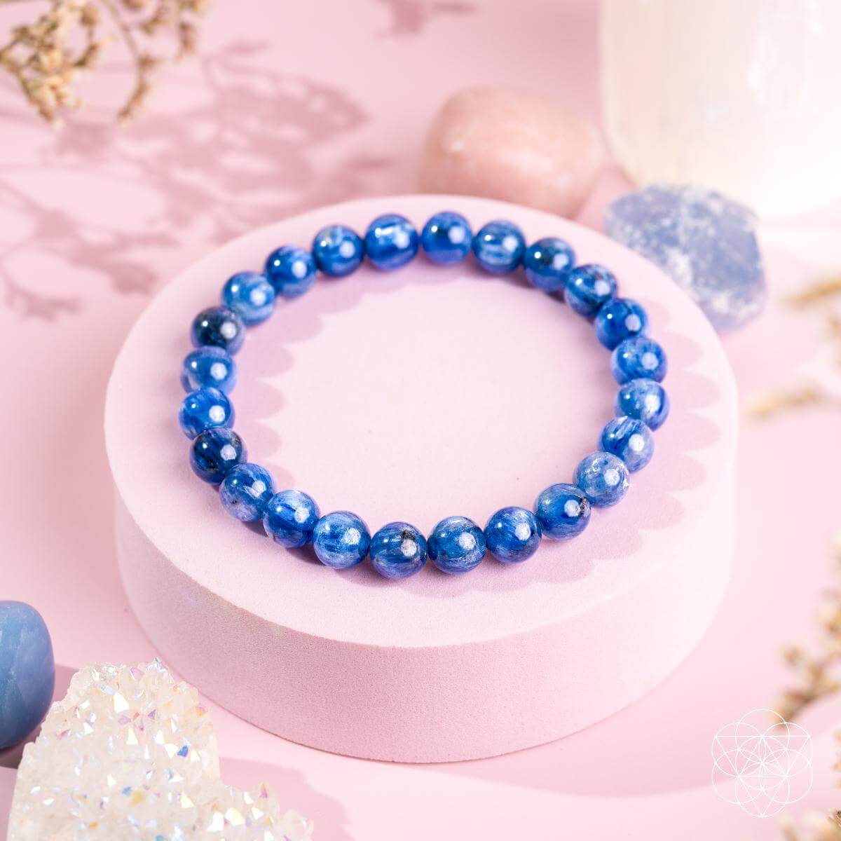 The My Truth, My Power – Rare Kyanite Bracelet by Conscious Items features shiny blue beads on a light pink circular stand, accented by pastel crystals and flowers—ideal for throat chakra balance against a soft pink background.