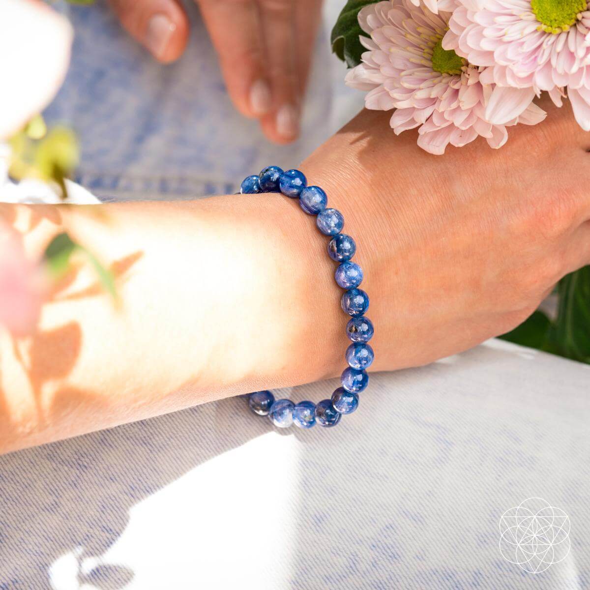 A hand adorned with the Conscious Items "My Truth, My Power – Rare Kyanite Bracelet" holds a bouquet of light pink flowers, sunlight casting gentle shadows on the wrist against a light background.