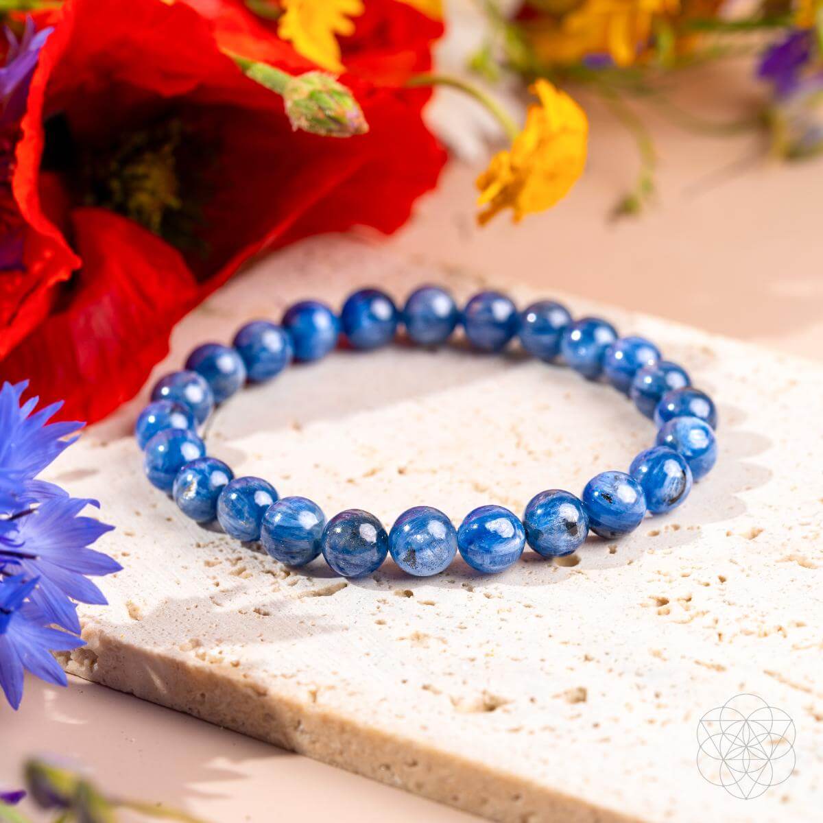 The My Truth, My Power – Rare Kyanite Bracelet by Conscious Items rests on a textured stone with colorful flowers, creating a bright, decorative scene that channels the soothing energy of the throat chakra.