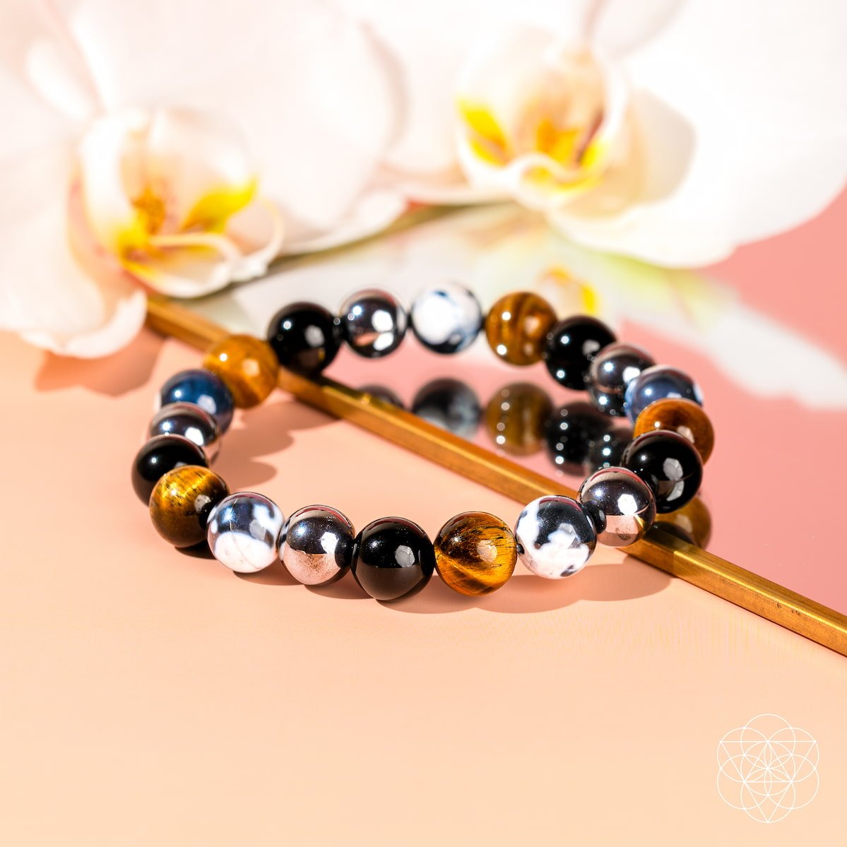 The Orca’s Armor - The Triple Protection & Healing Bracelet by Conscious Items features polished Orca Agate beads in brown, black, and white, displayed on a mirror with orchids—ideal for emotional healing.