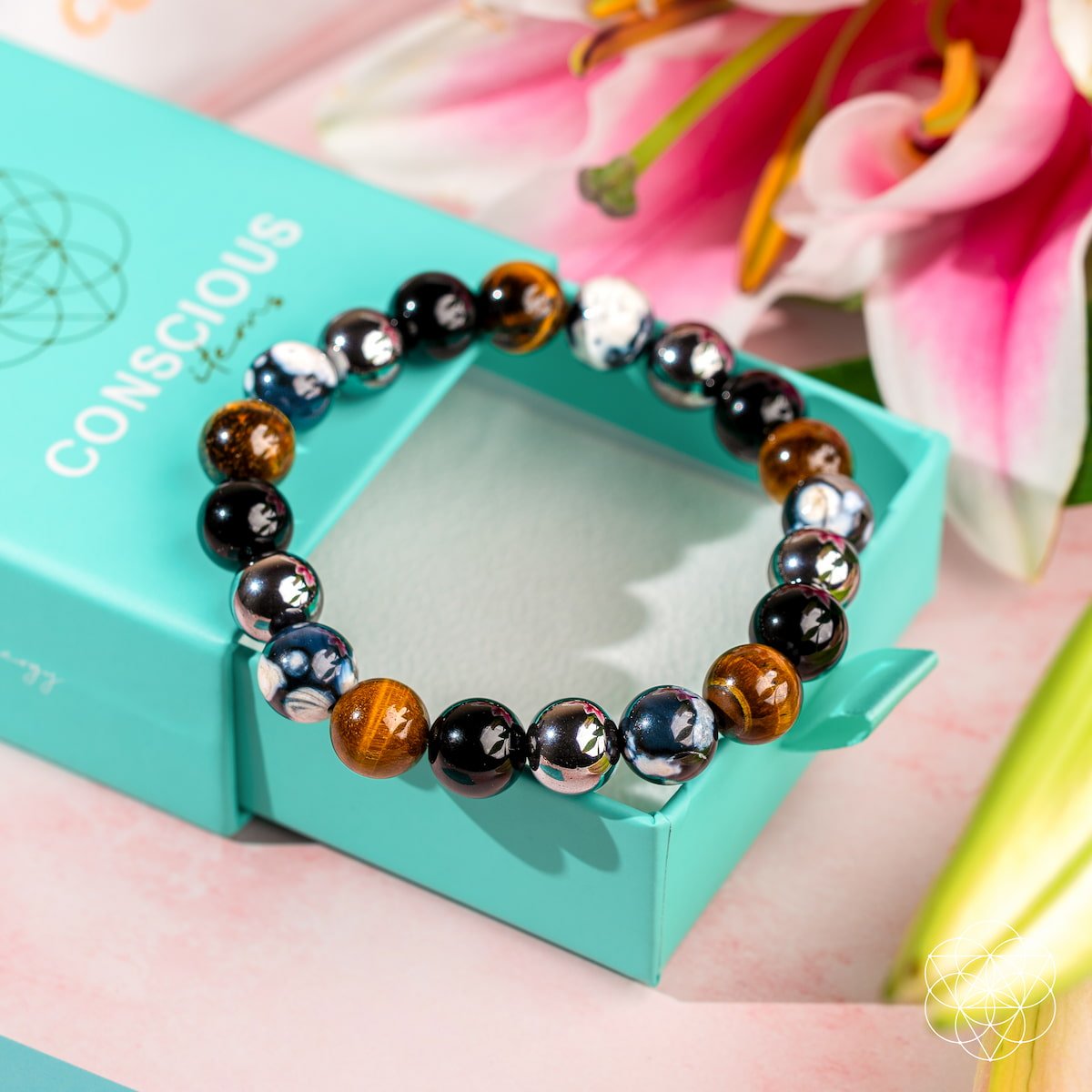 The Orca’s Armor - The Triple Protection & Healing Bracelet by Conscious Items, featuring black, brown, and marbled white-blue Orca Agate stones, is shown on a light turquoise box with pink lilies and green leaves in the background.