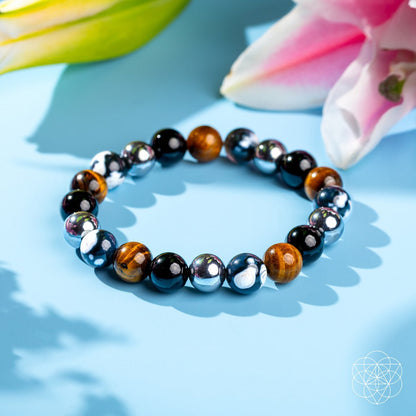 Orca’s Armor - The Triple Protection & Healing Bracelet by Conscious Items, featuring shiny black, silver, and brown tiger’s eye stones for emotional healing, is shown on a light blue surface with pink lilies and green leaves in the background.