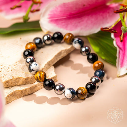 Orca’s Armor - The Triple Protection & Healing Bracelet by Conscious Items, crafted with glossy black, white, and brown Orca Agate for emotional healing, rests on beige stone slabs among pink lilies and green leaves.