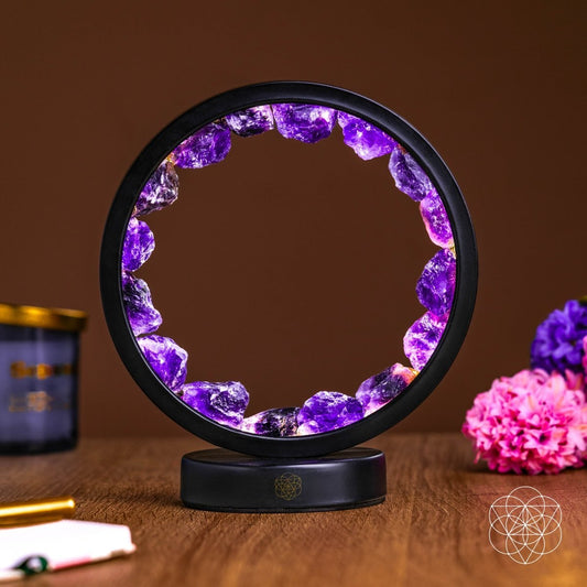 The Peace & Purification Duo by Conscious Items—a circular black amethyst lamp with glowing purple crystals—rests on a wooden surface, with blurred purple and pink flowers and a candle against a brown wall in the background.