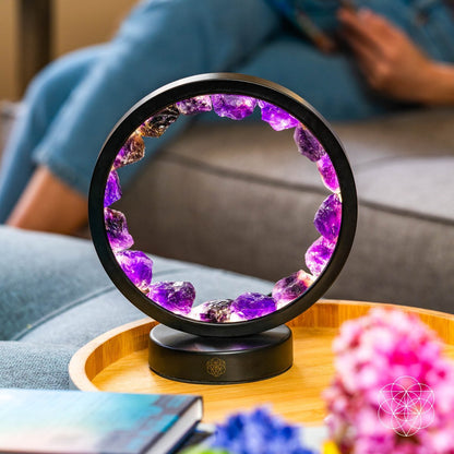 The Peace & Purification Duo by Conscious Items, featuring a circular amethyst lamp with purple crystals, rests on a wooden tray atop a gray couch. Pink and blue flowers are in the foreground; in the blurred background, someone in jeans holds a book.