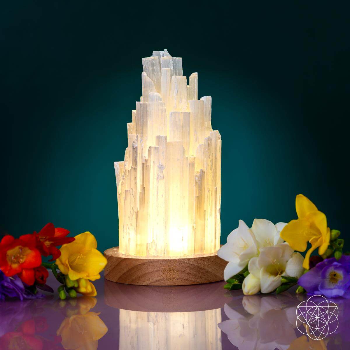 The Peace & Purification Duo by Conscious Items glows on a wooden base, surrounded by red, purple, yellow, and white flowers against teal. A geometric logo in the bottom right highlights the elegance of these crystal lamps.