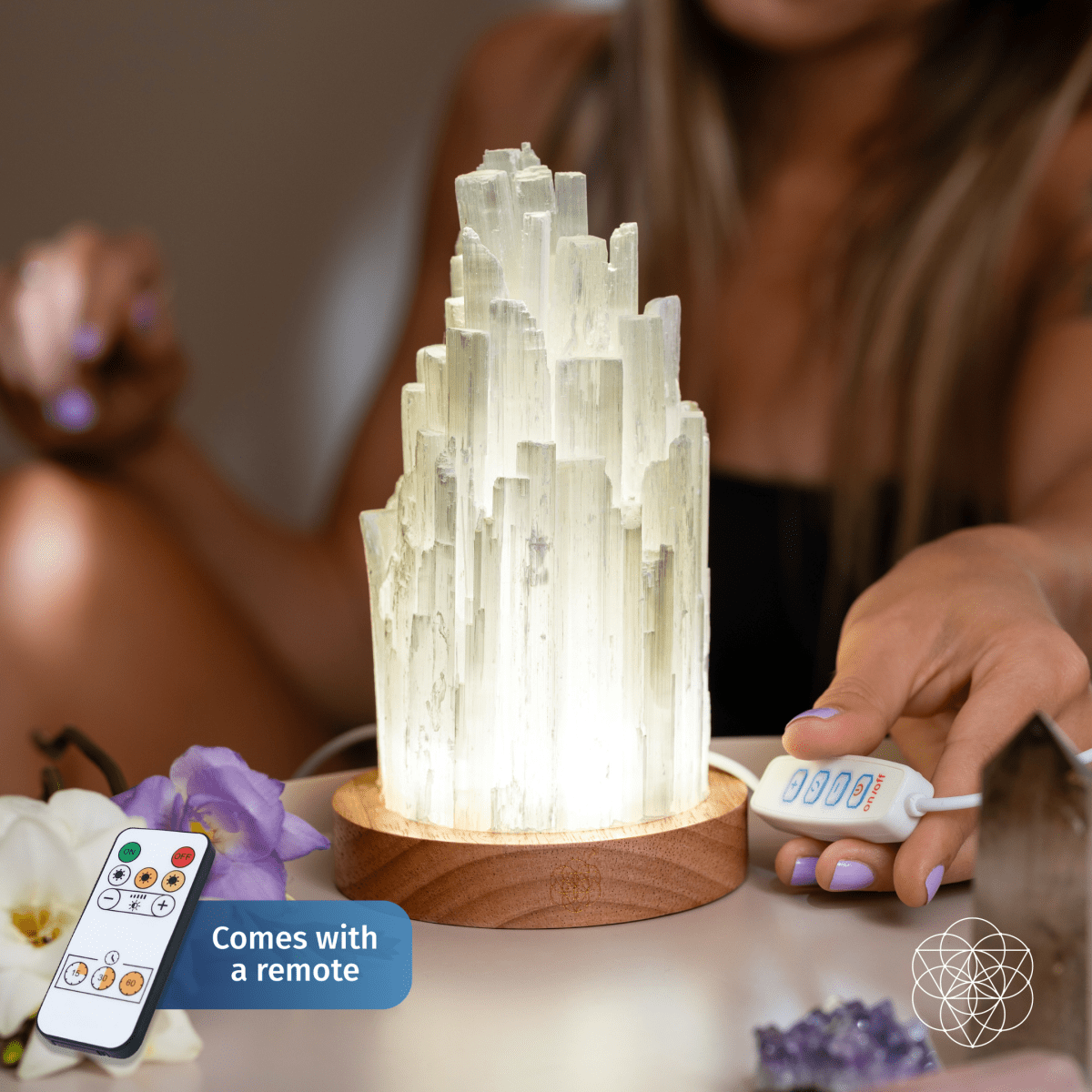 The Peace & Purification Duo by Conscious Items sits on a wooden base with a glowing selenite lamp. A hand holds its remote control, and another remote appears in the corner labeled “Comes with a remote.” Flowers and other crystal lamps are nearby.