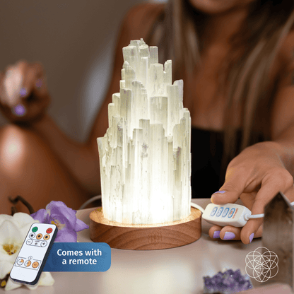 The Peace & Purification Duo by Conscious Items sits on a wooden base with a glowing selenite lamp. A hand holds its remote control, and another remote appears in the corner labeled “Comes with a remote.” Flowers and other crystal lamps are nearby.