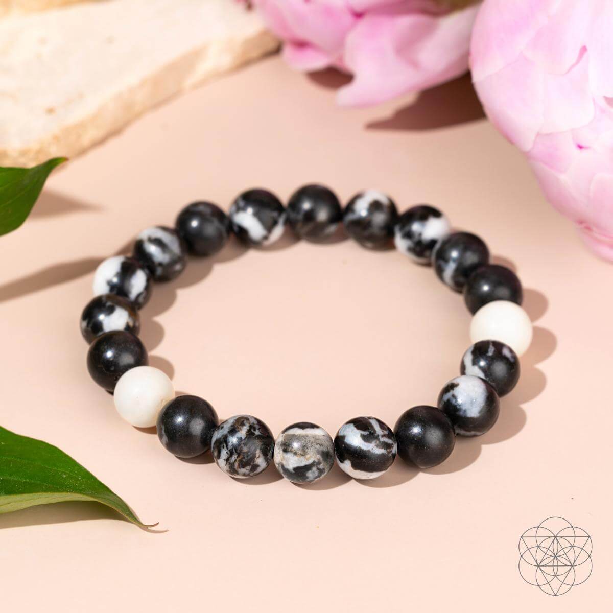 The Permission to Pause - The Zebra Jasper Bracelet by Conscious Items features black, white, and marbled Zebra Jasper beads for emotional balance, showcased on a light pink background with green leaves and pink flowers.