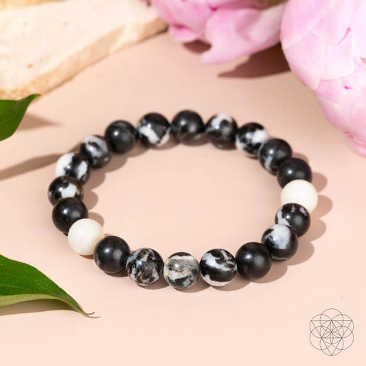 The Permission to Pause - The Zebra Jasper Bracelet by Conscious Items features black, white, and marbled Zebra Jasper beads for emotional balance, showcased on a light pink background with green leaves and pink flowers.