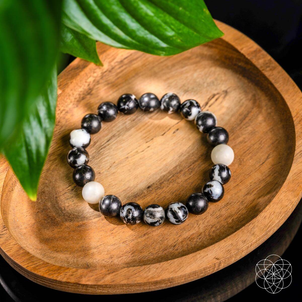 The "Permission to Pause - The Zebra Jasper Bracelet" by Conscious Items, featuring black and white marbled stones for emotional balance and burnout relief, rests on a wooden dish with green leaves in the corner and a geometric logo at the bottom right.