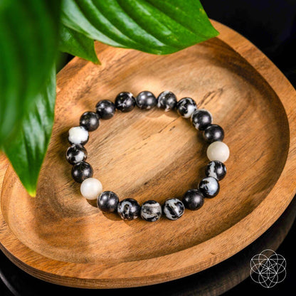 The "Permission to Pause - The Zebra Jasper Bracelet" by Conscious Items, featuring black and white marbled stones for emotional balance and burnout relief, rests on a wooden dish with green leaves in the corner and a geometric logo at the bottom right.