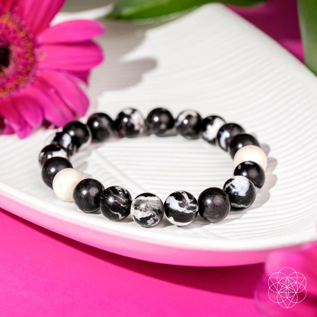 Permission to Pause - The Zebra Jasper Bracelet from Conscious Items rests on a white dish with a pink flower and green leaves, set against a bright pink background—ideal for emotional balance and burnout relief.