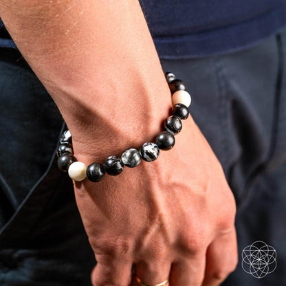 A close-up of a wrist wearing Conscious Items’ Permission to Pause - The Zebra Jasper Bracelet, featuring black, white, and marbled beads. The person wears a navy shirt and dark pants. A geometric logo appears in the bottom right corner.