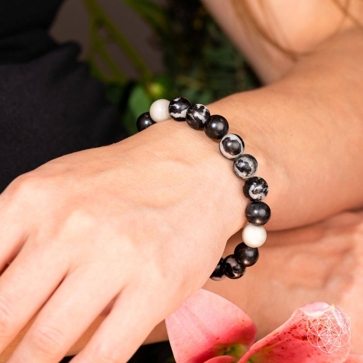 A close-up of a wrist wearing Conscious Items' Permission to Pause - The Zebra Jasper Bracelet, featuring black, white, and marbled beads. The hand rests on a pink flower amid greenery, symbolizing emotional balance and burnout relief.