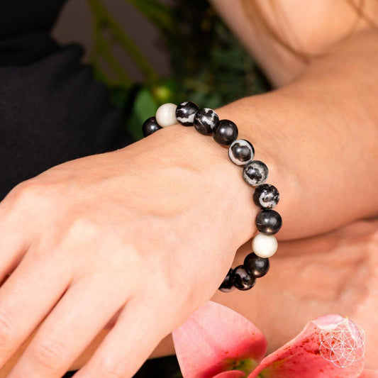 A close-up of a wrist wearing Conscious Items' Permission to Pause - The Zebra Jasper Bracelet, featuring black, white, and marbled beads. The hand rests on a pink flower amid greenery, symbolizing emotional balance and burnout relief.