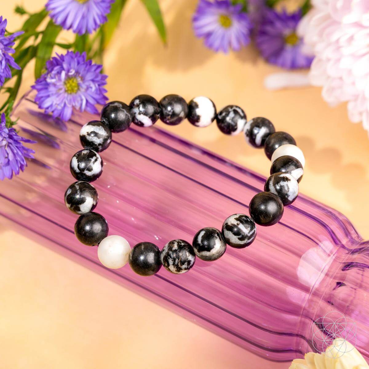 Permission to Pause - The Zebra Jasper Bracelet by Conscious Items, known for burnout relief and emotional balance, is showcased on a translucent purple ribbed glass bottle amid purple and white flowers on a soft peach background.