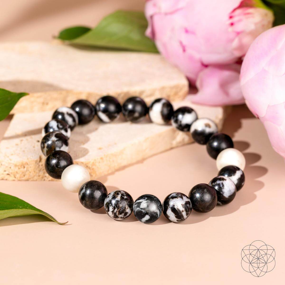 The Permission to Pause - The Zebra Jasper Bracelet by Conscious Items features black, white, and marbled beads, displayed on a light surface with green leaves and pink flowers—perfect for encouraging emotional balance.