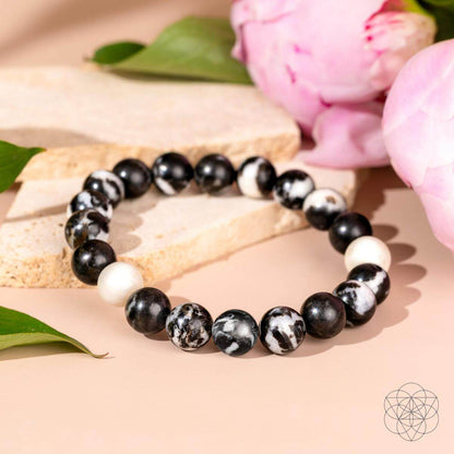 The Permission to Pause - The Zebra Jasper Bracelet by Conscious Items features black, white, and marbled beads, displayed on a light surface with green leaves and pink flowers—perfect for encouraging emotional balance.