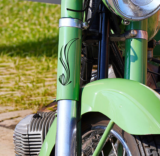 Close-up of the front part of a green motorcycle with a black decorative design on the fork, a round headlight, and grassy background visible.