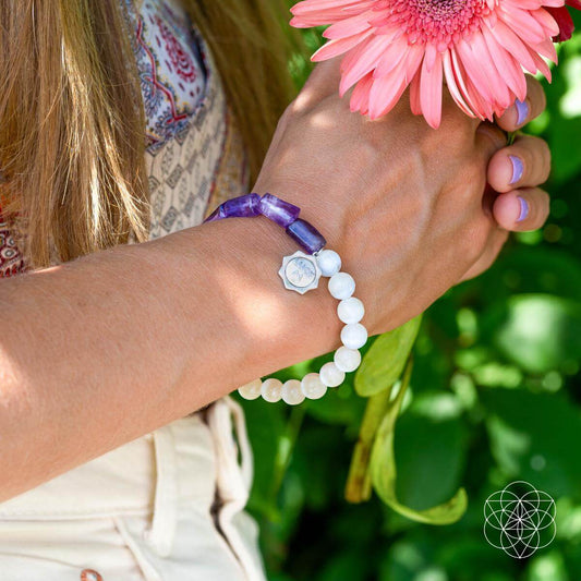 A person holding a pink flower wears the Pure Luck - Horseshoe Amethyst Bracelet by Conscious Items, featuring white and purple stones, a selenite crystal, and a silver charm. They’re dressed in light colors with a leafy green background.