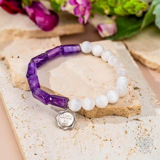The Pure Luck - Horseshoe Amethyst Bracelet by Conscious Items, with purple and white beads and a silver LUCK horseshoe charm, is shown on a textured stone surface with green and purple flowers behind it.