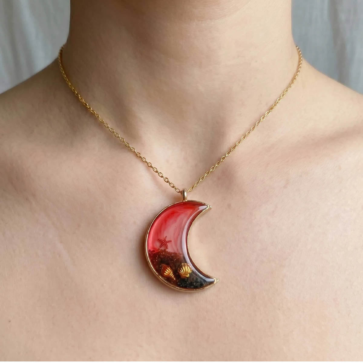 A crescent moon-shaped pendant with a gold metal frame filled with red and white marbled resin, black volcanic-style sand, and gold seashell and starfish charms.