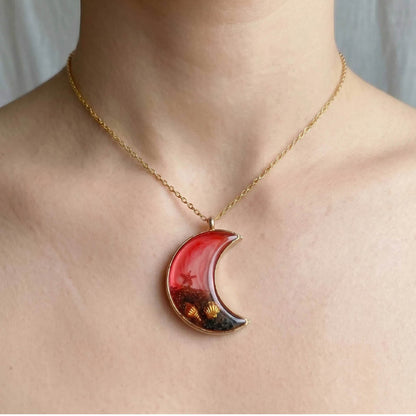 A crescent moon-shaped pendant with a gold metal frame filled with red and white marbled resin, black volcanic-style sand, and gold seashell and starfish charms.