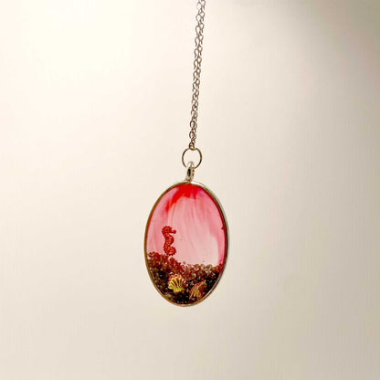 An oval silver-framed pendant containing a red and clear marbled resin background, dark coarse sand at the base, and small metallic gold and copper charms of a seahorse and seashells.