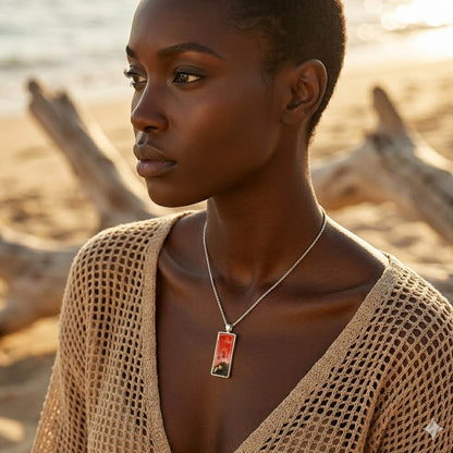 A person with dark skin standing on a beach, wearing a rectangular silver pendant on a chain. The pendant contains a gradient of translucent red resin over a dark, textured base with small gold nautical charms.