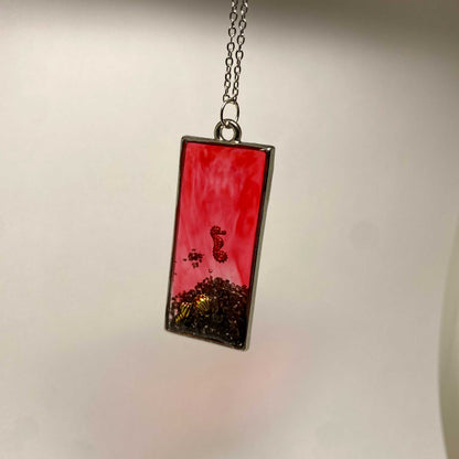Rectangular pendant with red and black design on a chain against a neutral background