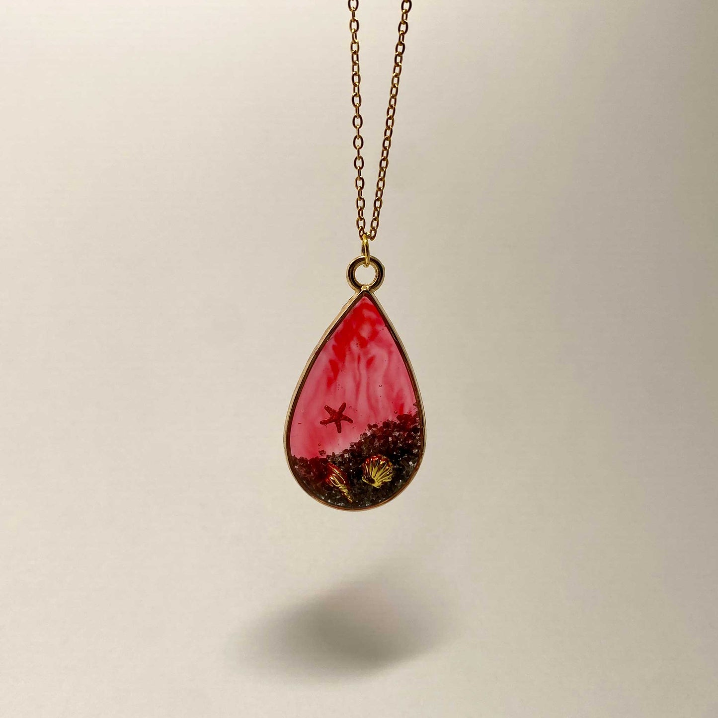 A teardrop-shaped gold-toned pendant filled with translucent red resin, containing a miniature red starfish and two small gold seashells resting on dark, textured sediment.