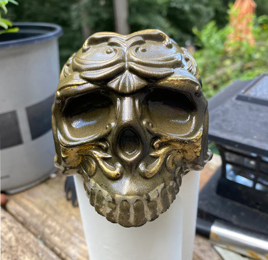 A decorative skull with ornate, leafy patterns and an antique bronze finish is displayed outdoors on a white surface, with plants and garden items blurred in the background.