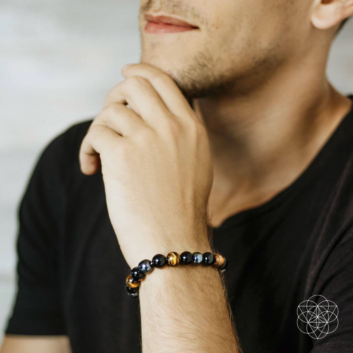 A man in a black t-shirt rests his chin on his hand, displaying the Conscious Items Sacred Healing & Protection Trio bracelet with shiny black and brown stones on his wrist.