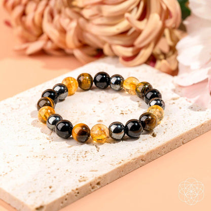 The Sacred Healing & Protection Trio by Conscious Items—a Black Friday exclusive bracelet with black, silver, and amber-yellow beads—is shown on a stone slab with soft-focus flowers in the background.