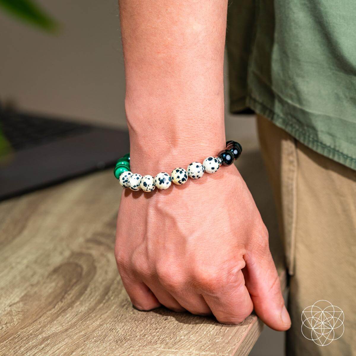A person in a green shirt and khaki pants wears the Soul Reset: Dalmatian Jasper Bracelet of Healing by Conscious Items—known to absorb negativity and support emotional healing—standing beside a wooden table with a softly blurred background.