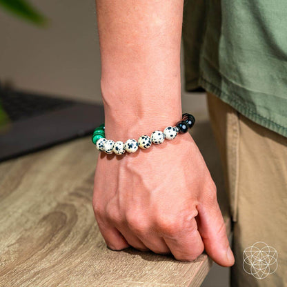 A person in a green shirt and khaki pants wears the Soul Reset: Dalmatian Jasper Bracelet of Healing by Conscious Items—known to absorb negativity and support emotional healing—standing beside a wooden table with a softly blurred background.