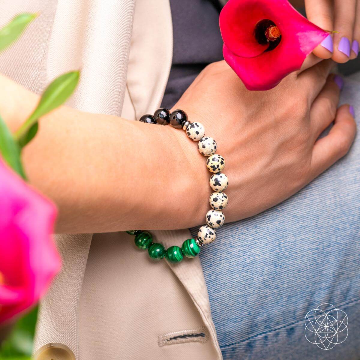 A person wearing the Soul Reset: Dalmatian Jasper Bracelet of Healing by Conscious Items holds a pink calla lily. Dressed in a beige blazer and jeans, they show off purple nail polish amid pink flowers in the foreground.