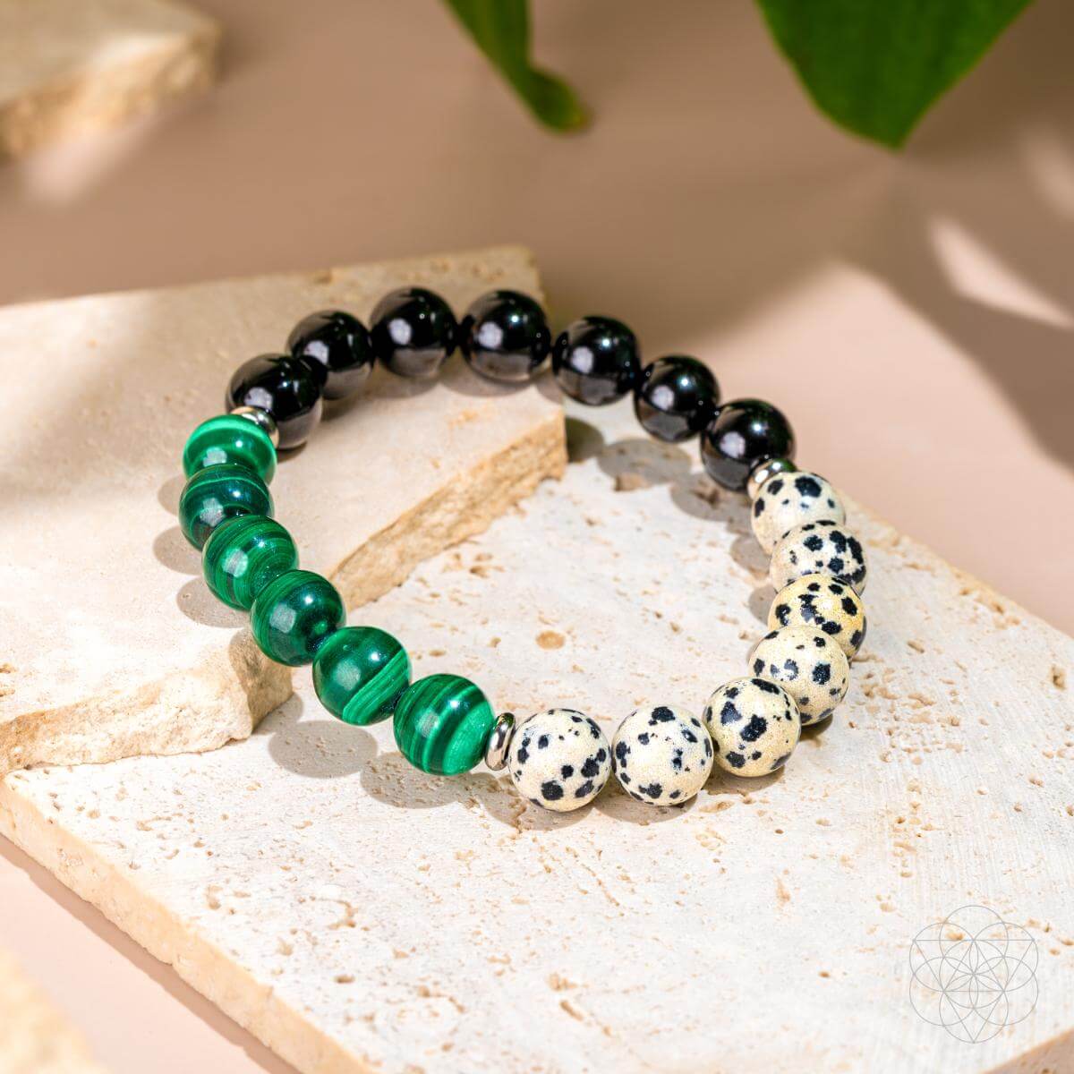 The Soul Reset: Dalmatian Jasper Bracelet of Healing by Conscious Items, featuring Dalmatian Jasper, shiny black, and striped green stones, is crafted to absorb negativity and support emotional healing. Shown on a textured beige surface under soft lighting.