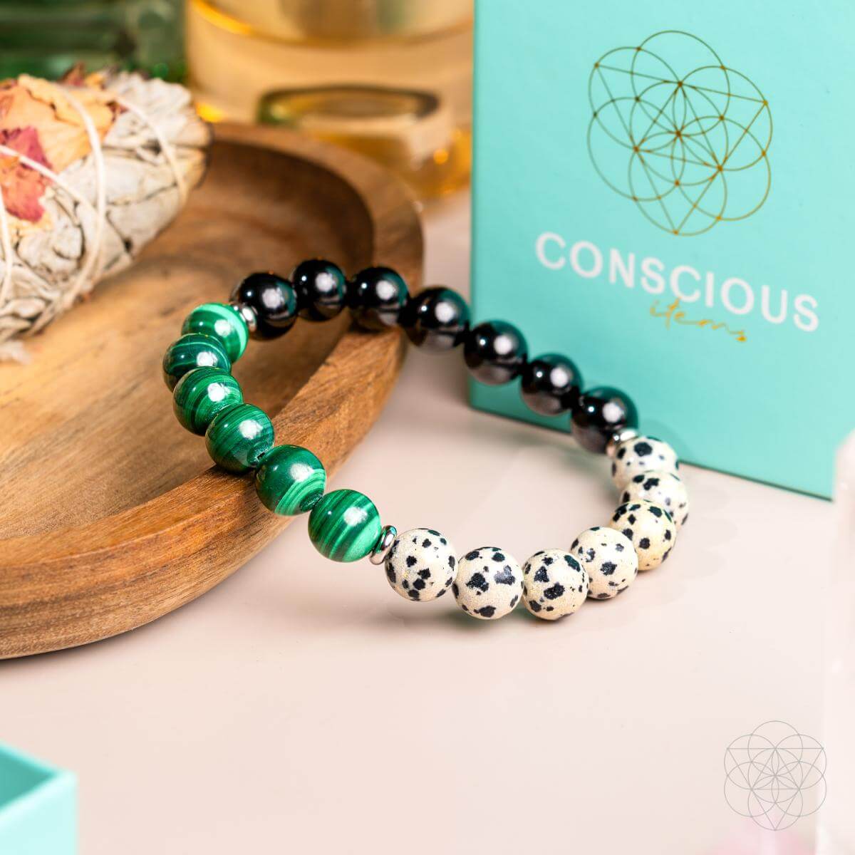 The Soul Reset: Dalmatian Jasper Bracelet of Healing by Conscious Items, featuring green malachite and black and white Dalmatian Jasper for emotional healing, sits on a white surface near a turquoise box, wooden tray, and sage.