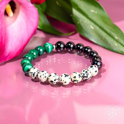 The Soul Reset: Dalmatian Jasper Bracelet of Healing by Conscious Items features black, green, and white beads, displayed on a glossy pink surface with a pink flower and green leaves, blending style with emotional healing.