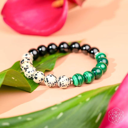The Soul Reset: Dalmatian Jasper Bracelet of Healing by Conscious Items, adorned with negativity-absorbing jasper and green striped beads, lays on a leaf amid pink petals against a peach background.