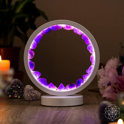 The Stress Less, Sleep More – Amethyst Healing Lamp by Conscious Items, featuring purple crystals, glows on a wooden table with candles, flowers, and heart-shaped accents nearby. Plants and softly lit décor create a calming atmosphere in the background.
