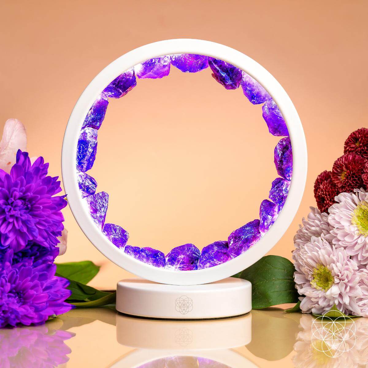 The Stress Less, Sleep More – Amethyst Healing Lamp by Conscious Items features glowing purple crystals in a white circular frame, surrounded by vibrant flowers, creating a calming oasis against a soft peach backdrop.