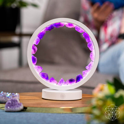 The Stress Less, Sleep More – Amethyst Healing Lamp by Conscious Items glows softly on a table, its purple crystals radiating calm. Blurred plants and a person in vibrant attire evoke the ambiance of a serene healing oasis.