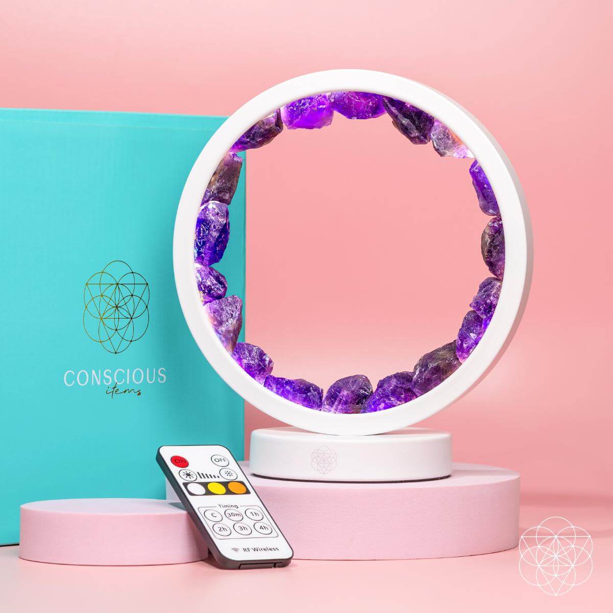 The Stress Less, Sleep More – Amethyst Healing Lamp by Conscious Items, a circular white lamp filled with purple crystals, is shown on pink platforms with its remote in front and a teal Conscious Items box behind—all on a pink backdrop.